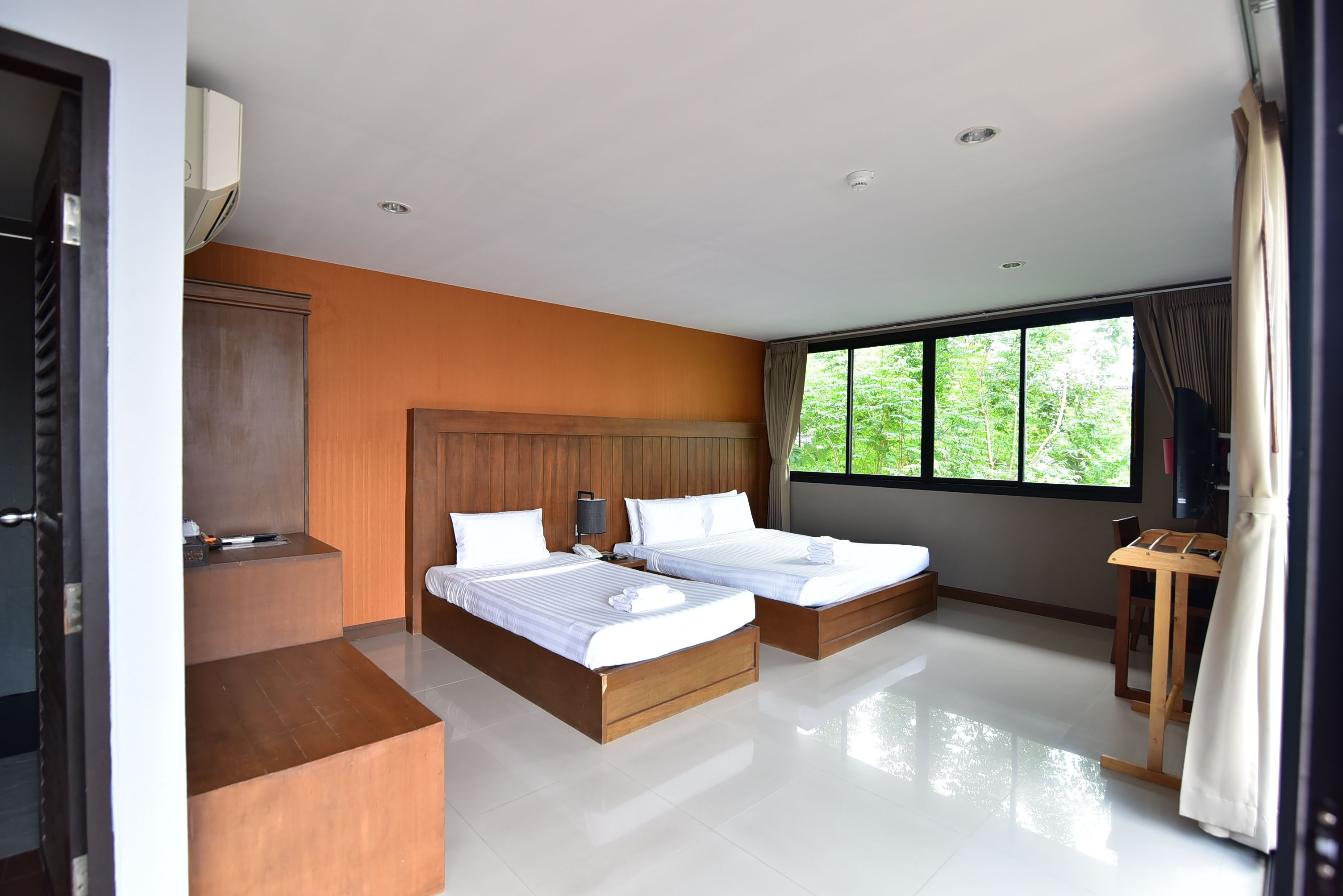 A modern hotel room with two beds, a wooden headboard, a desk, and a large window offering a view of greenery.