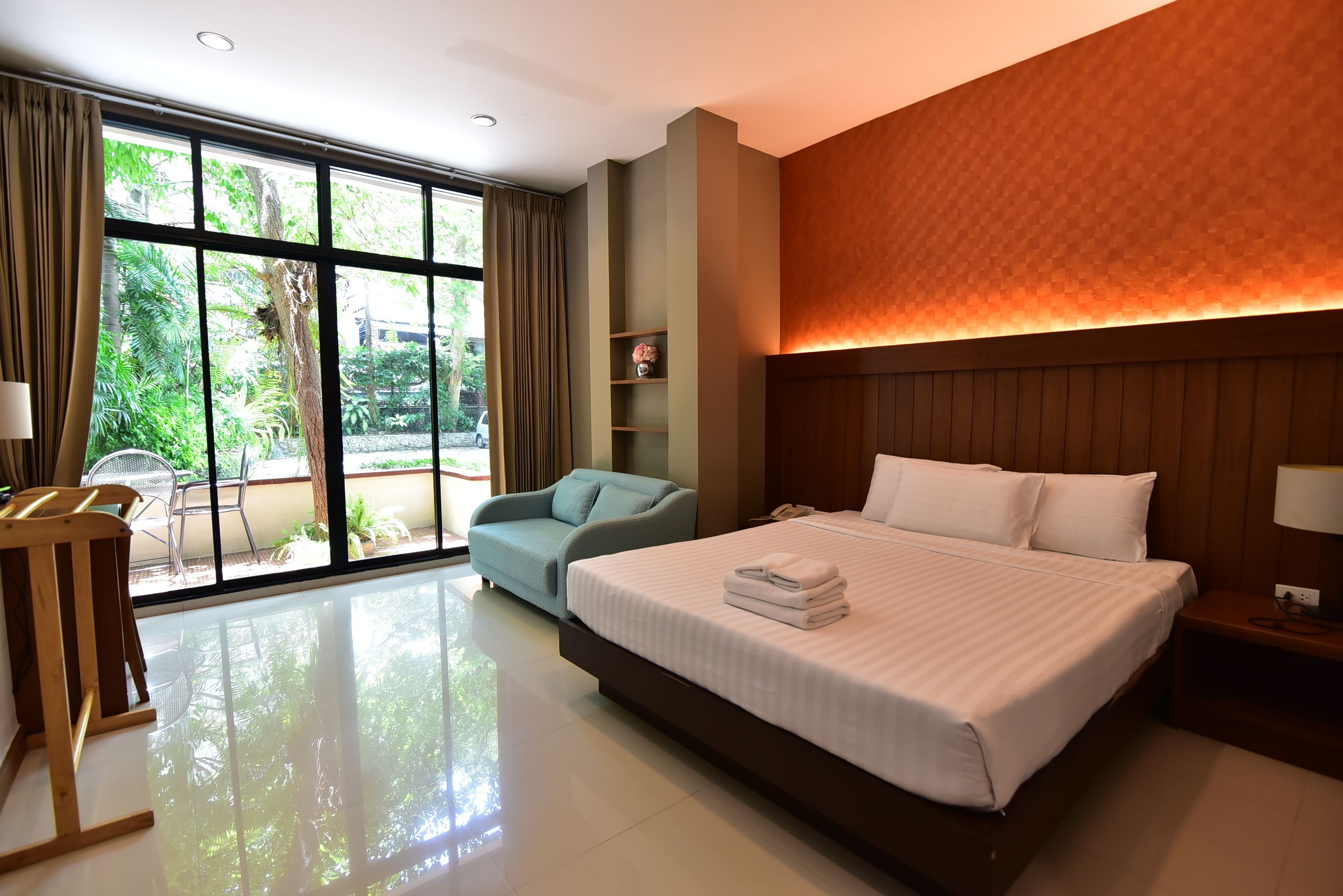 A hotel room with a large bed, a sitting area with a chair, a small table, and a view of the outdoors through large windows.
