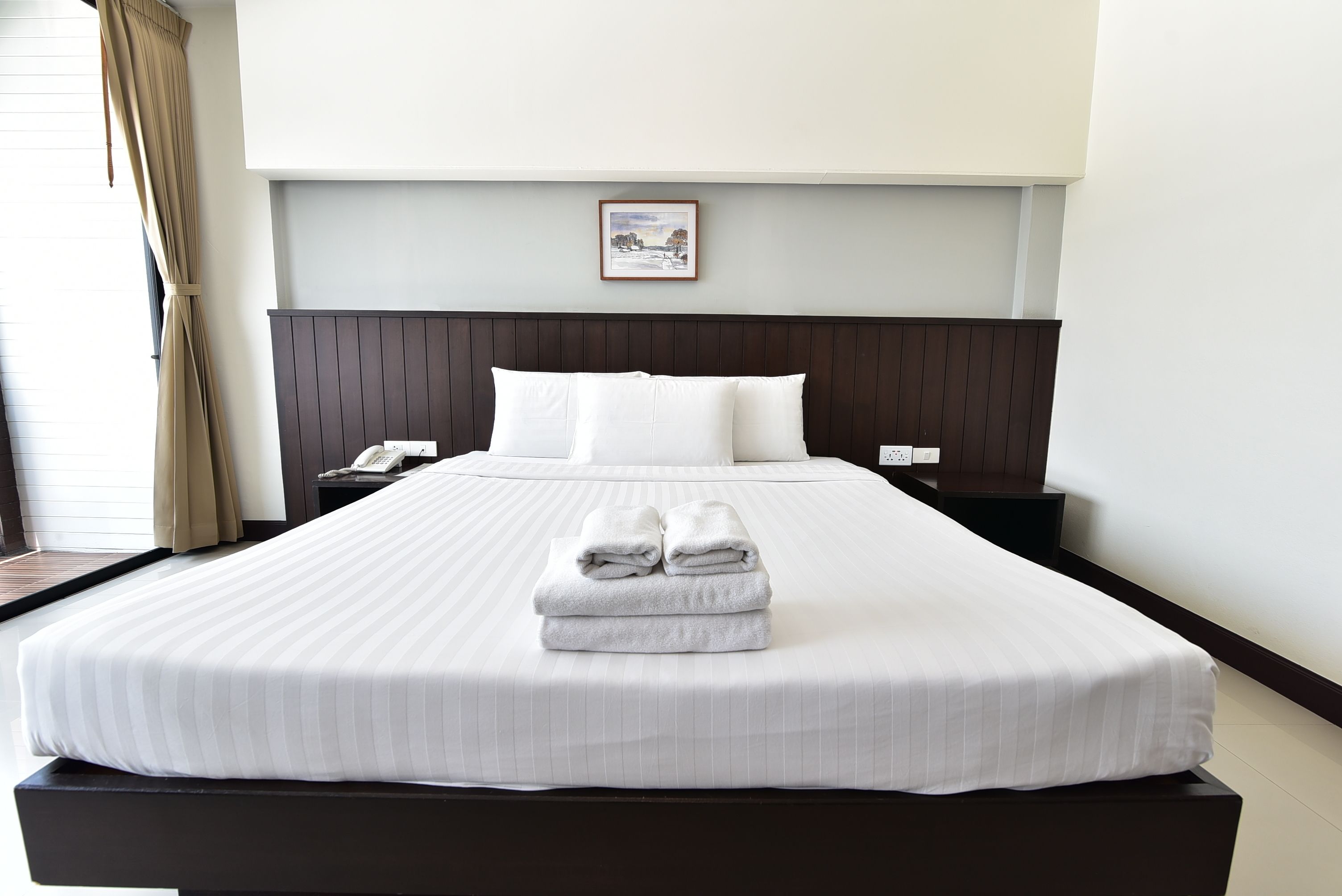 Una cama bien tendida con sábanas blancas y dos toallas dobladas, un cabecero de madera oscura, una mesita de noche con un teléfono y un cuadro en la pared.