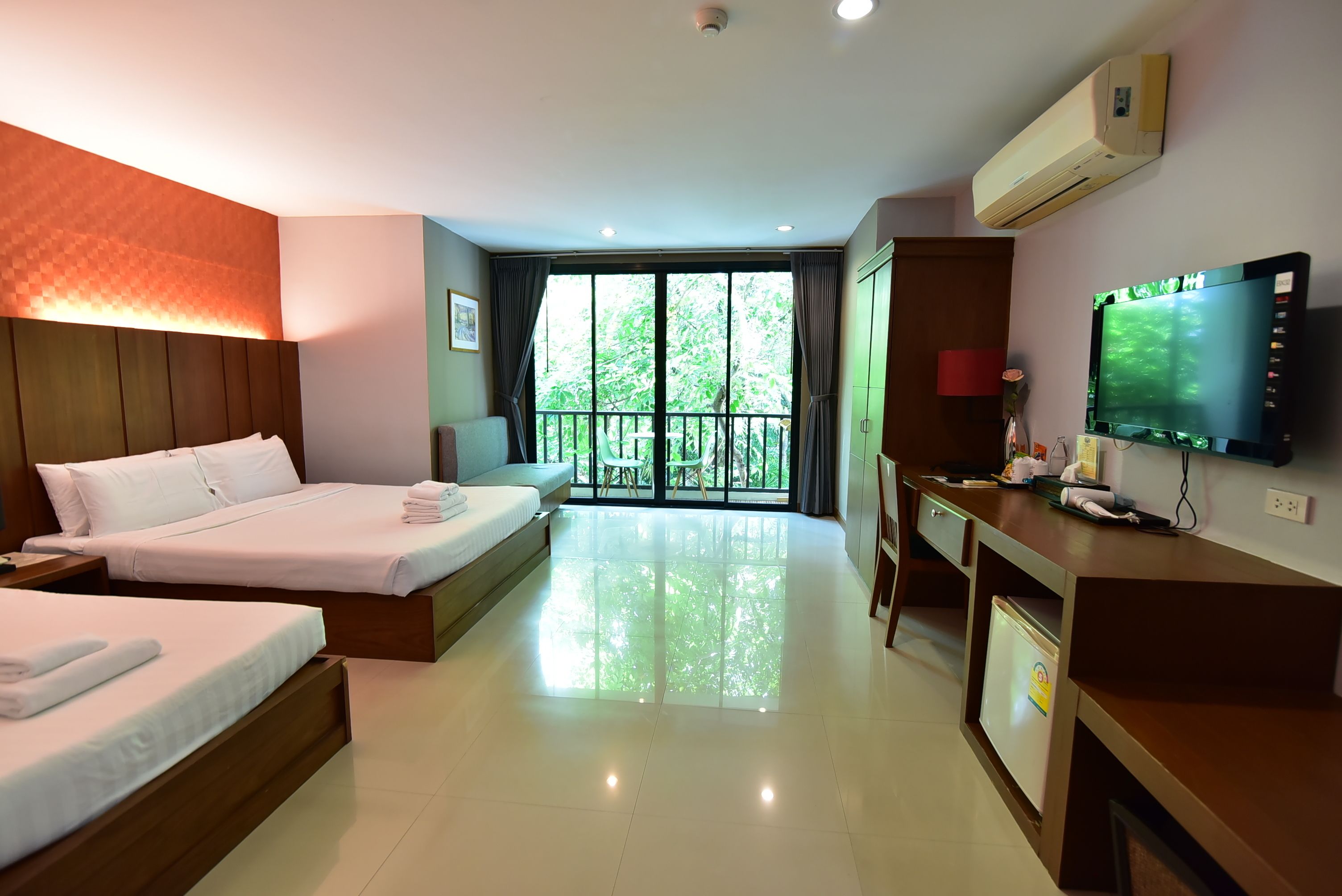 A hotel room with two beds, a desk, a television, and a balcony with a view of trees.