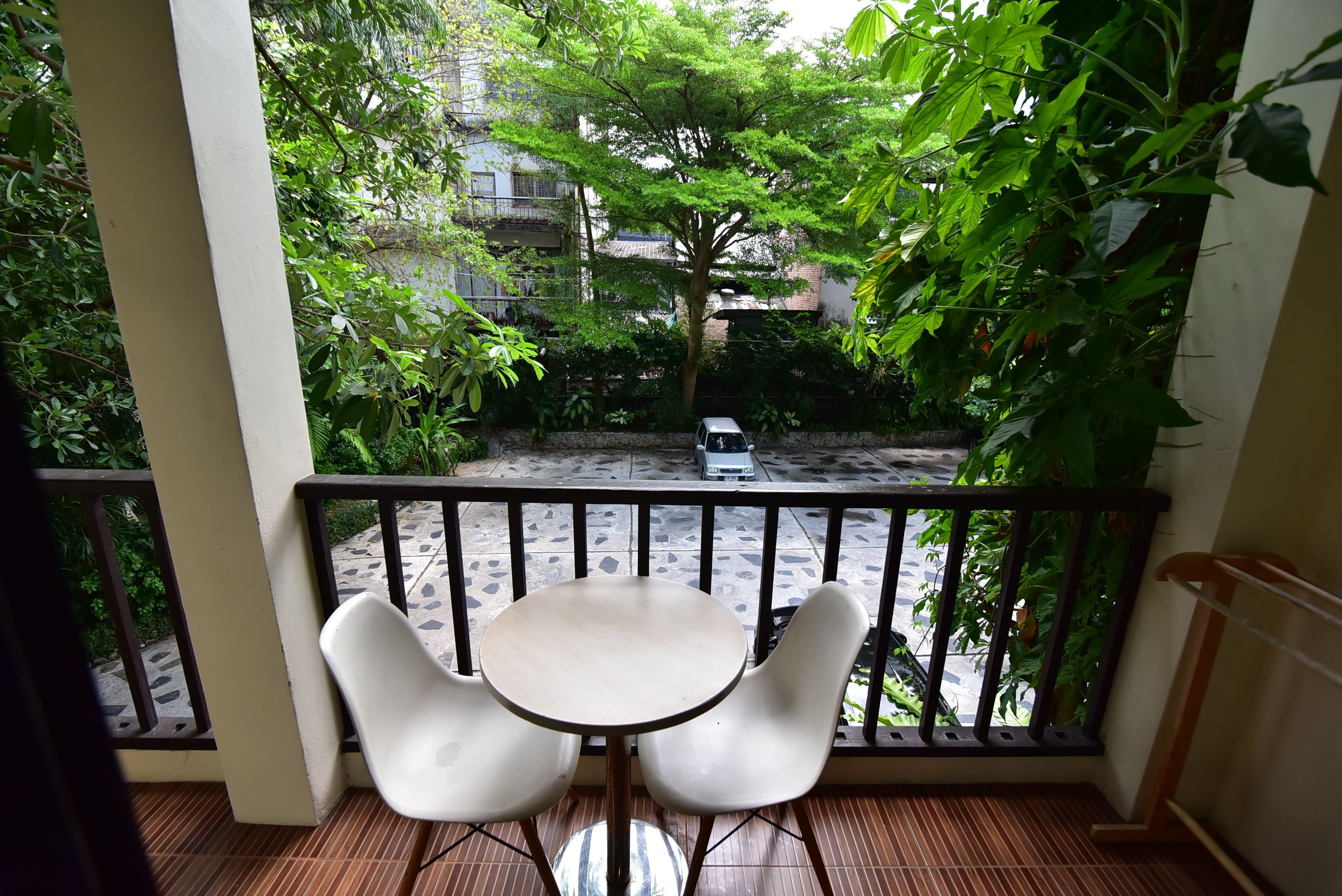 A balcony with a small table and two chairs, overlooking a garden with a tree and a parked car.