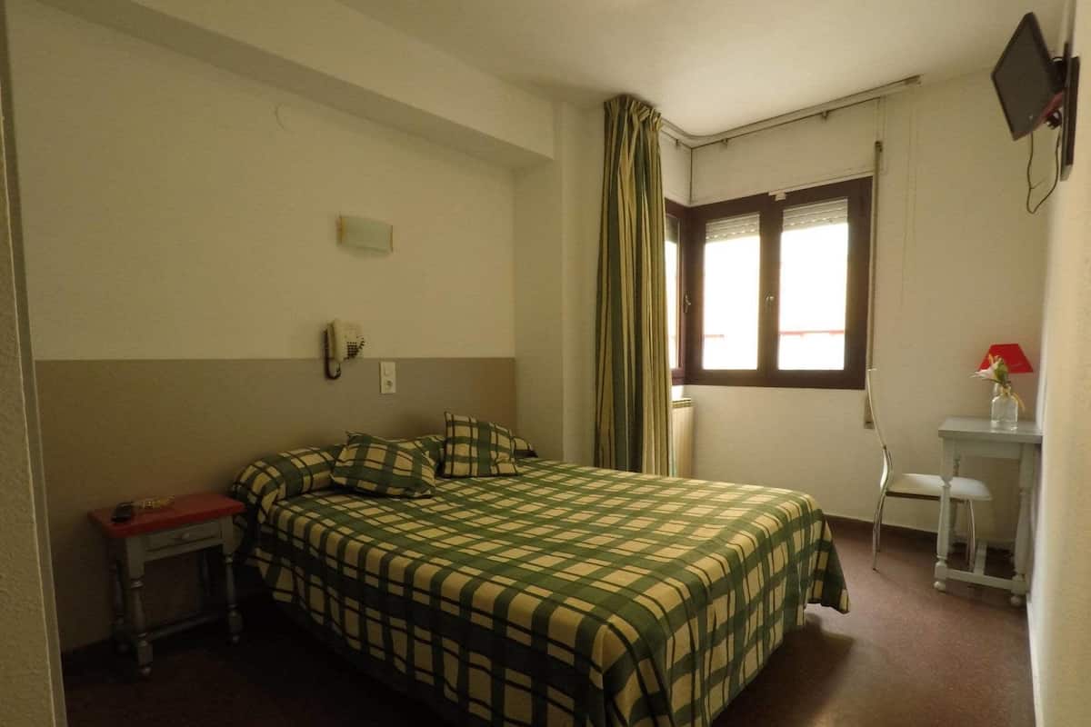 A single-bed room with a checkered bedspread, a bedside table with a lamp, a chair, and a desk with a computer monitor.