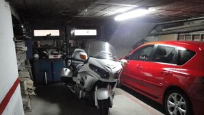 A motorcycle and a red car parked in a garage with a concrete floor and various tools and equipment in the background. - Hostal Palafox (Barbastro)