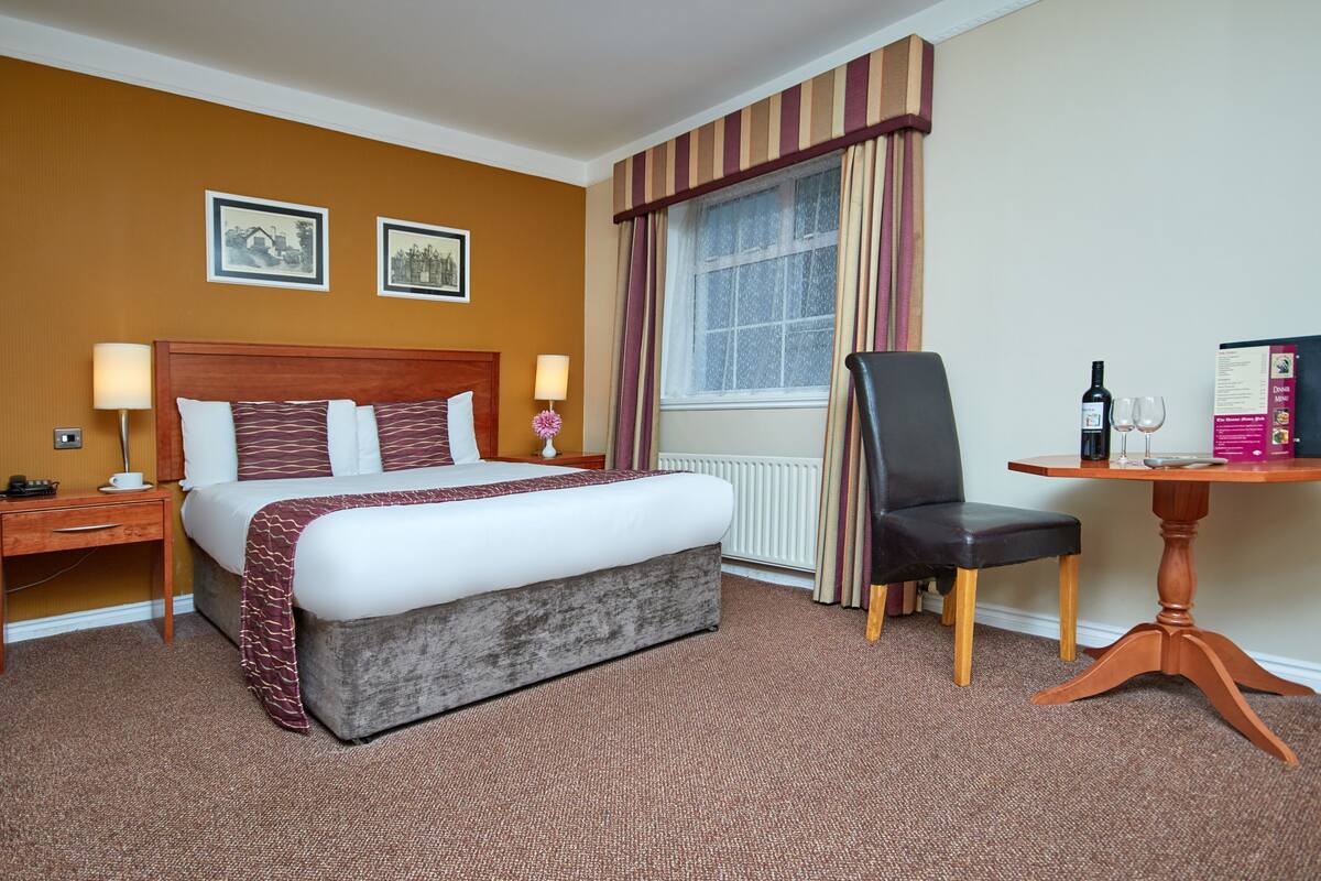A hotel room with a large bed, a wooden nightstand, a desk with a chair, a window with curtains, and a small table with a wine bottle and glasses.