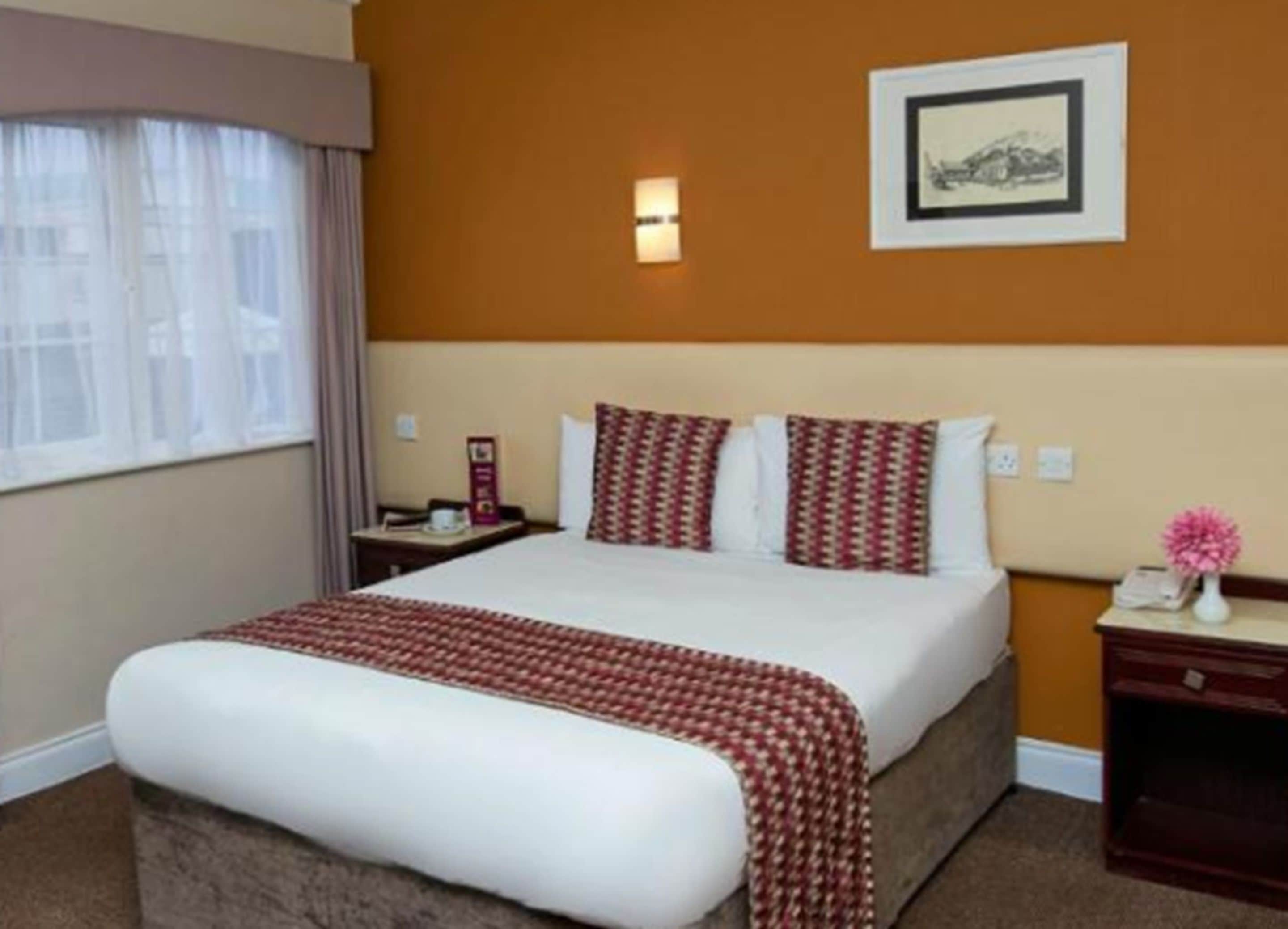 A hotel room with a large bed, a nightstand, a bedside table, a framed picture on the wall, and a window with curtains.