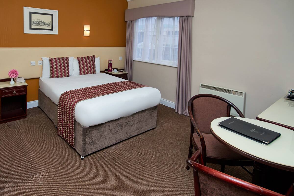 A hotel room with a bed, bedside tables, a chair, a desk, a window with curtains, and a wall-mounted picture.