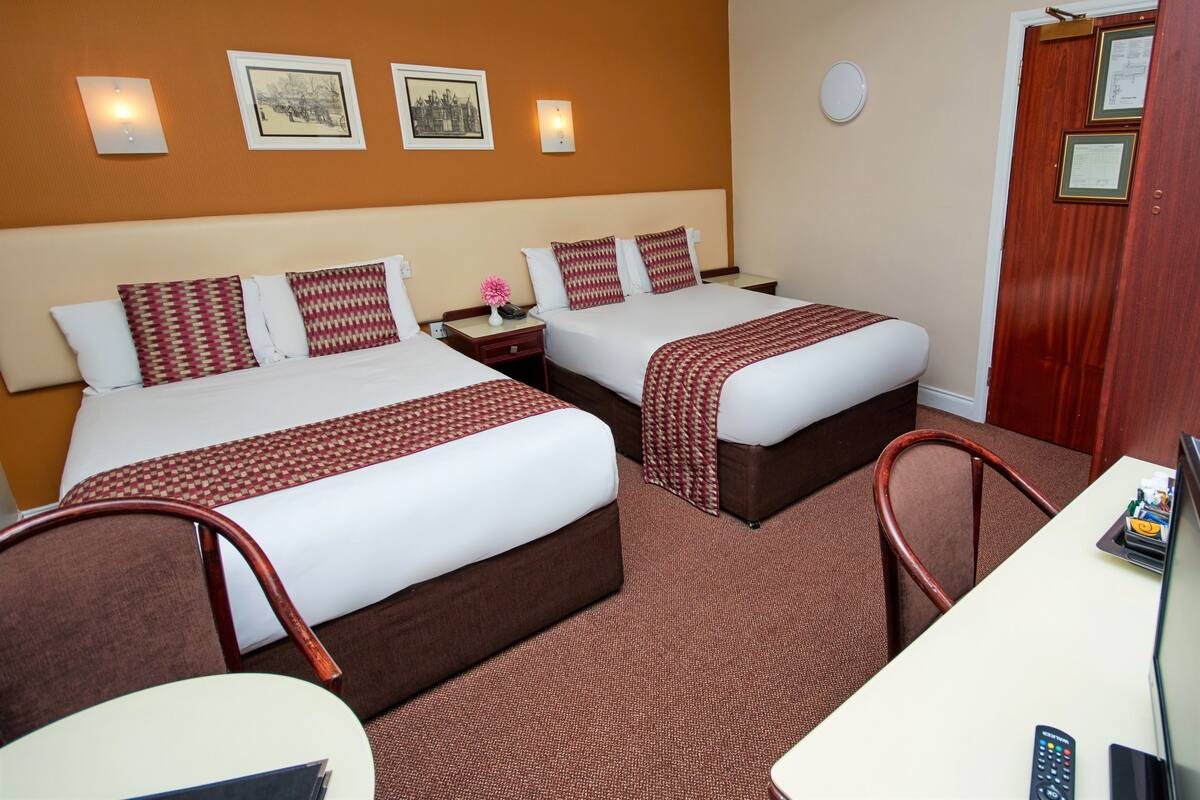 A hotel room with two beds, a desk, and a chair. There are framed pictures on the wall, a clock, and a door with a plaque.
