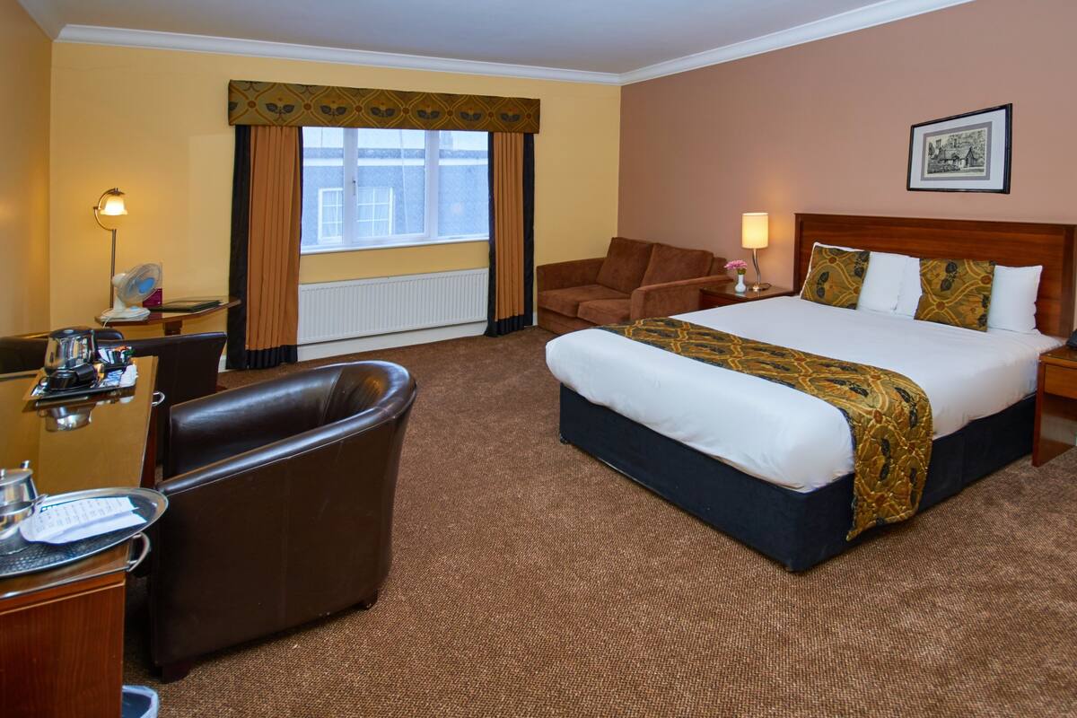 A hotel room with a large bed, a desk with a chair, a sofa, and a window with curtains.