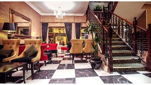 Eine Lobby mit Schachbrettboden, einer Treppe mit aufwendiger Geländerarbeiten und einer Sitzecke mit Sesseln und einer Couch.