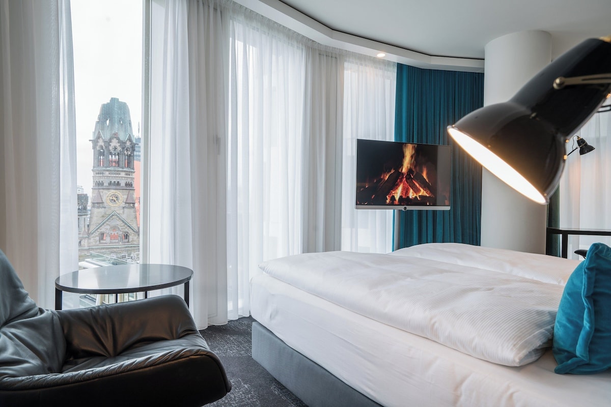 A hotel room with a bed, a desk, a chair, a TV, and a view of a cityscape.
