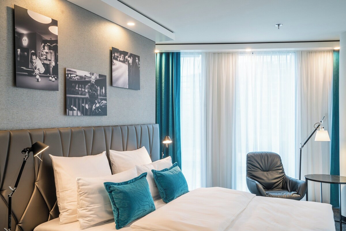 A modern hotel room with a large bed, a headboard with built-in lighting, a seating area with a chair and table, and a large window with curtains.