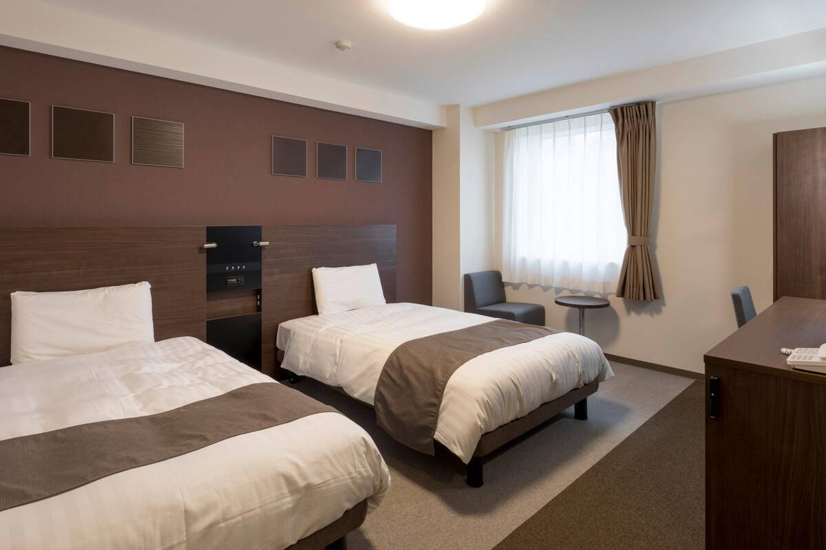 A hotel room with two beds, a desk, a chair, a small table, and a window with curtains.