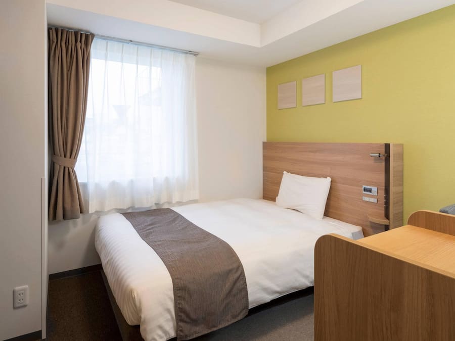 A hotel room with a bed, a desk, a window with curtains, and a wall with three pictures.