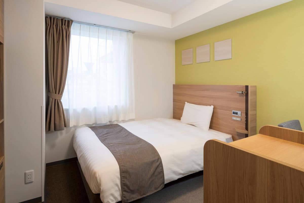 A hotel room with a bed, a desk, a window with curtains, and a wall with three framed pictures.