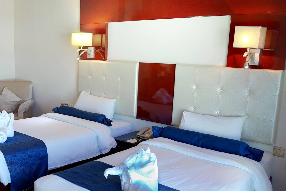 A hotel room with two beds, each with a blue bedspread and white linens. There are two bedside lamps, a chair, and a red accent wall.