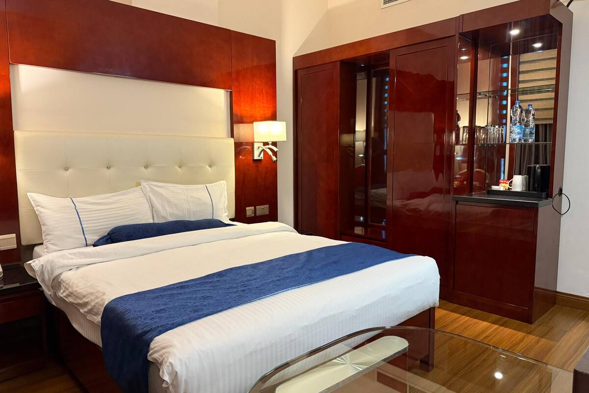 A hotel room with a large bed, a wooden wardrobe, a glass table, and a mirror.