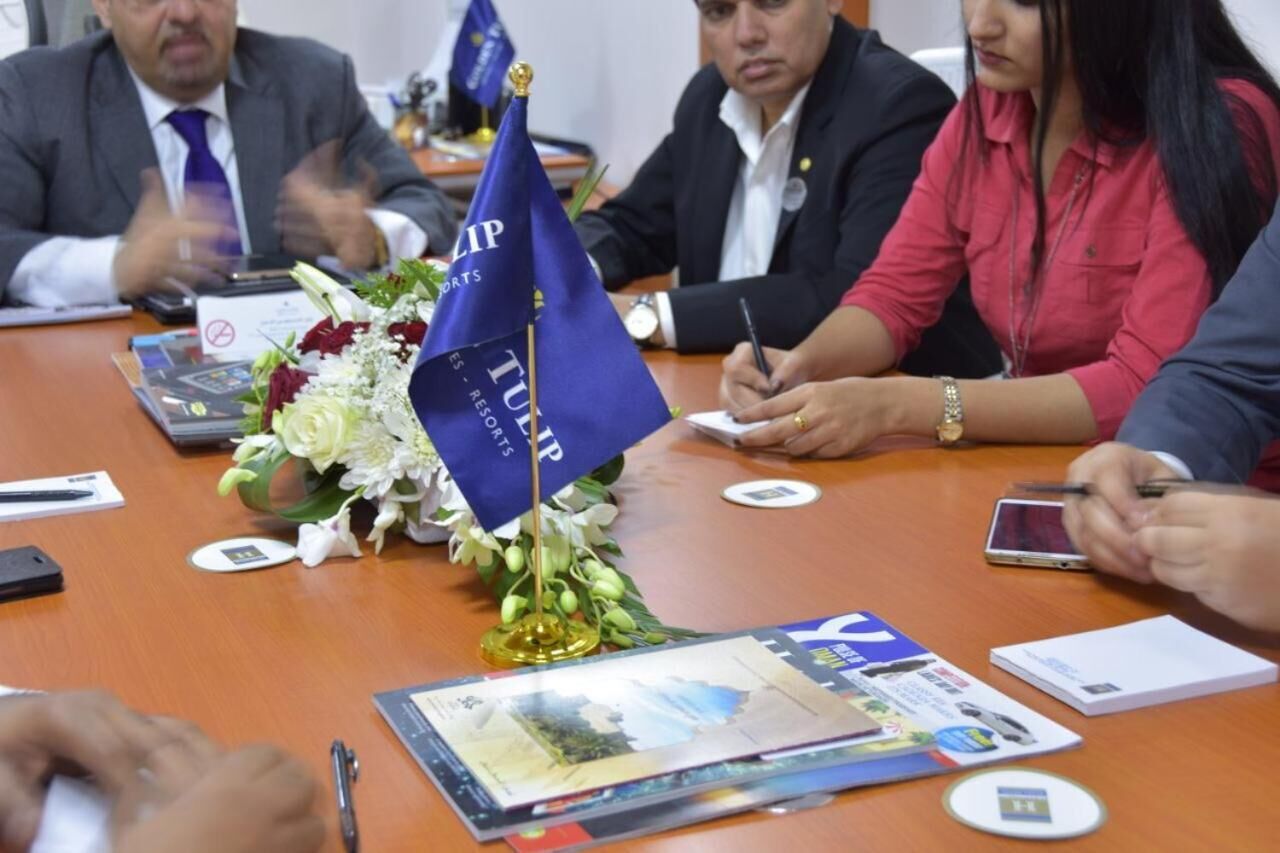 Uma reunião com participantes ao redor de uma mesa, com uma bandeira, documentos e um arranjo floral.