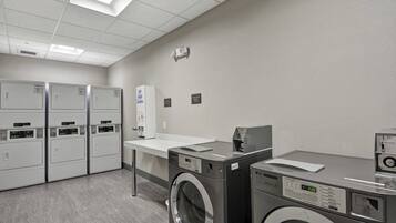 A laundry facility with multiple washing machines and dryers, a white counter, and a wall-mounted control panel.