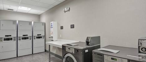 Une buanderie équipée de plusieurs machines à laver et de plusieurs sèche-linge, d’un comptoir blanc et d’un panneau de commande fixé au mur.