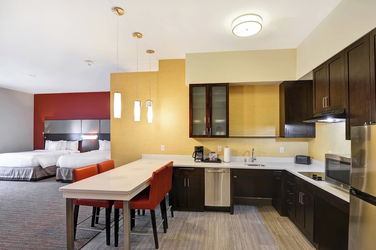 A modern hotel room with a separate dining area, a bed with a headboard, a kitchen with dark cabinets, a refrigerator, and a coffee maker.