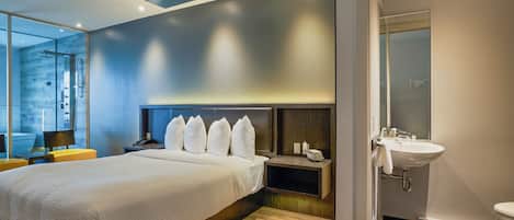 A modern hotel room with a large bed, bedside tables, a nightstand, a bathroom with a sink and toilet, and a view of the city.