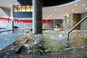 An indoor swimming pool with a hot tub area, a diving platform, and a person in the water.