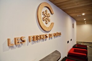 Ein Empfangsbereich mit einem hölzernen Schild, das 'LES FERMES DE CHATEAU' zeigt, und einem Logo darüber. Es gibt rote Sitzbereiche und eine Holzbalkendecke.