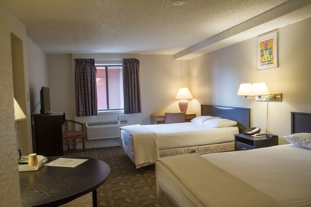 A hotel room with two beds, a desk, a chair, a TV, a window with curtains, and a painting on the wall.
