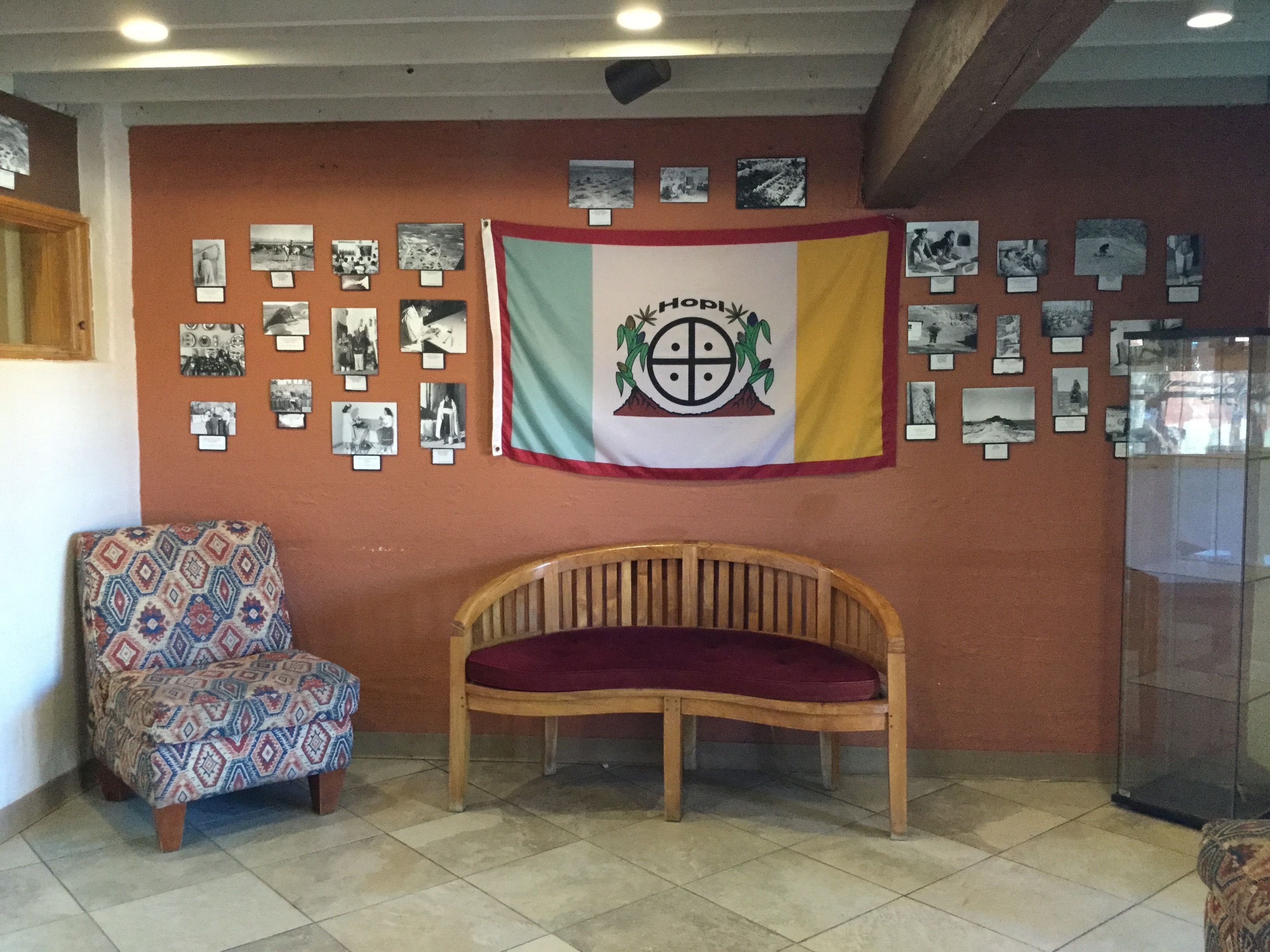 Una habitación con un banco de madera, un sillón estampado y una pared decorada con fotografías y una bandera.