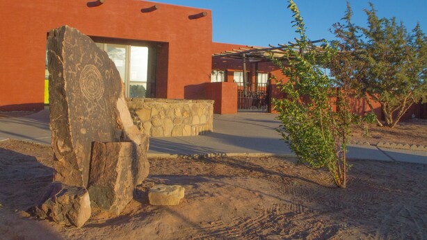 A building with a modern design, featuring a large rock sculpture in the foreground, a stone wall, and a small tree. - Hopi Cultural Center (Second Mesa)