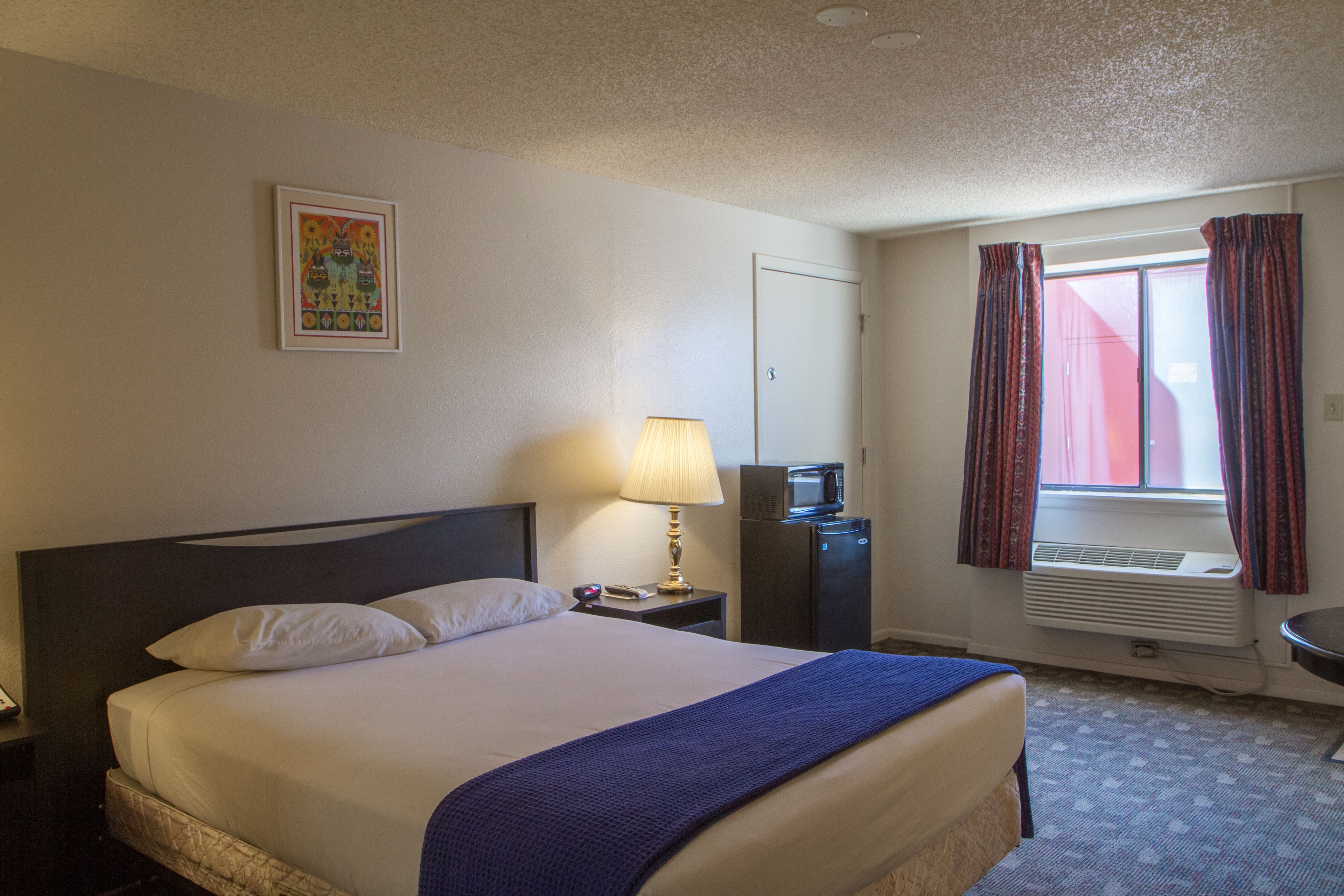 A hotel room with a bed, a bedside lamp, a desk with a computer, a window with curtains, and a wall-mounted air conditioner.
