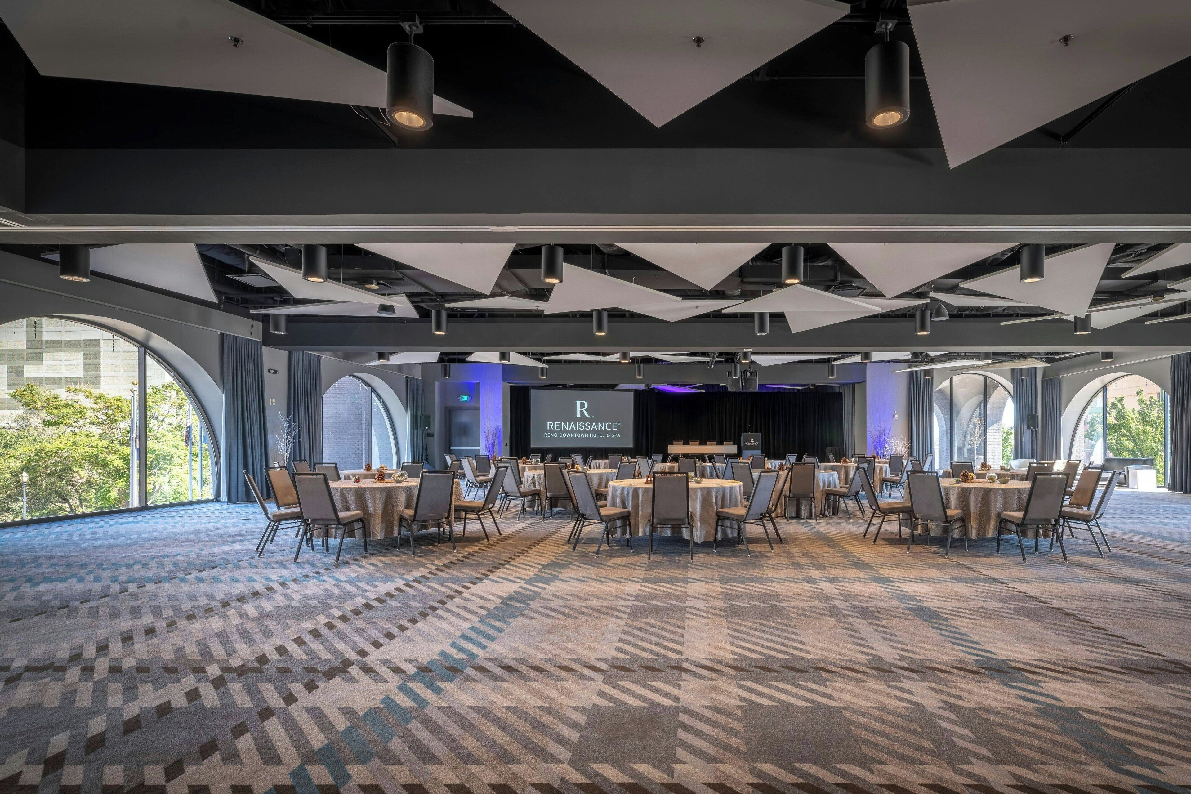 A spacious conference room with a patterned carpet, multiple tables with chairs, and large windows offering a view of the outdoors.