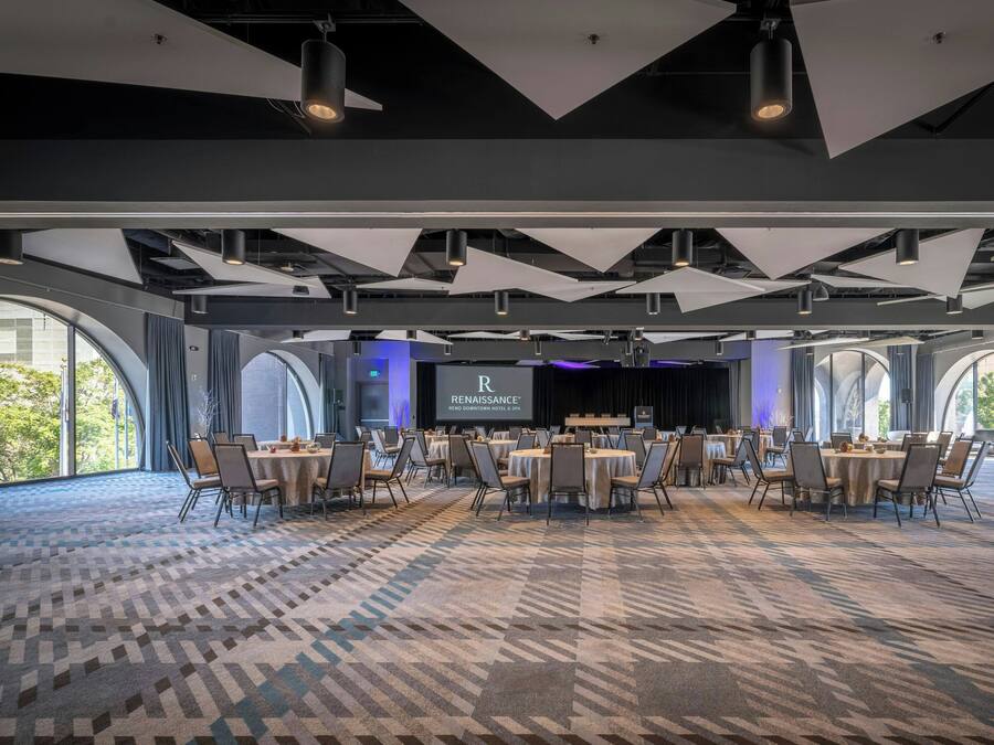 A spacious conference room with a patterned carpet, multiple tables with chairs, and large windows offering a view of the outdoors.