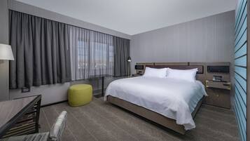 A modern hotel room with a large bed, a desk with a lamp, a small yellow ottoman, and a view of the city through the window.