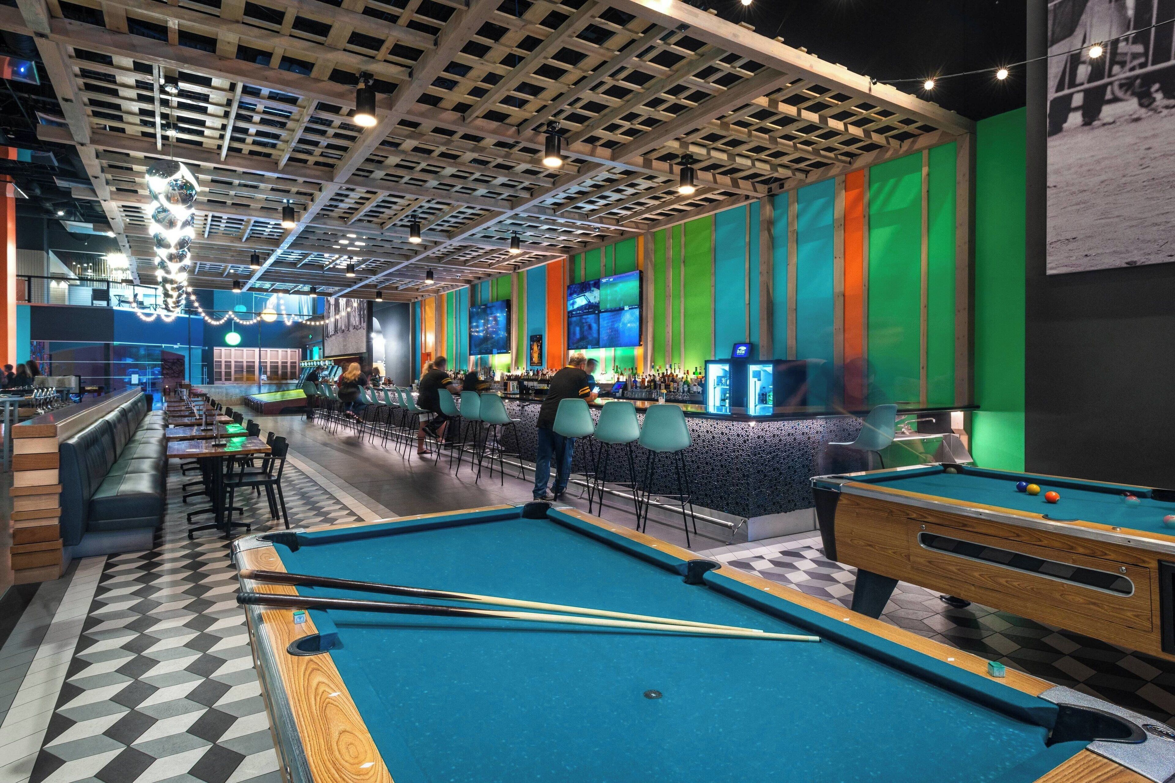 A pool table area with a checkered floor, a bar with seating, and a colorful wall with screens.