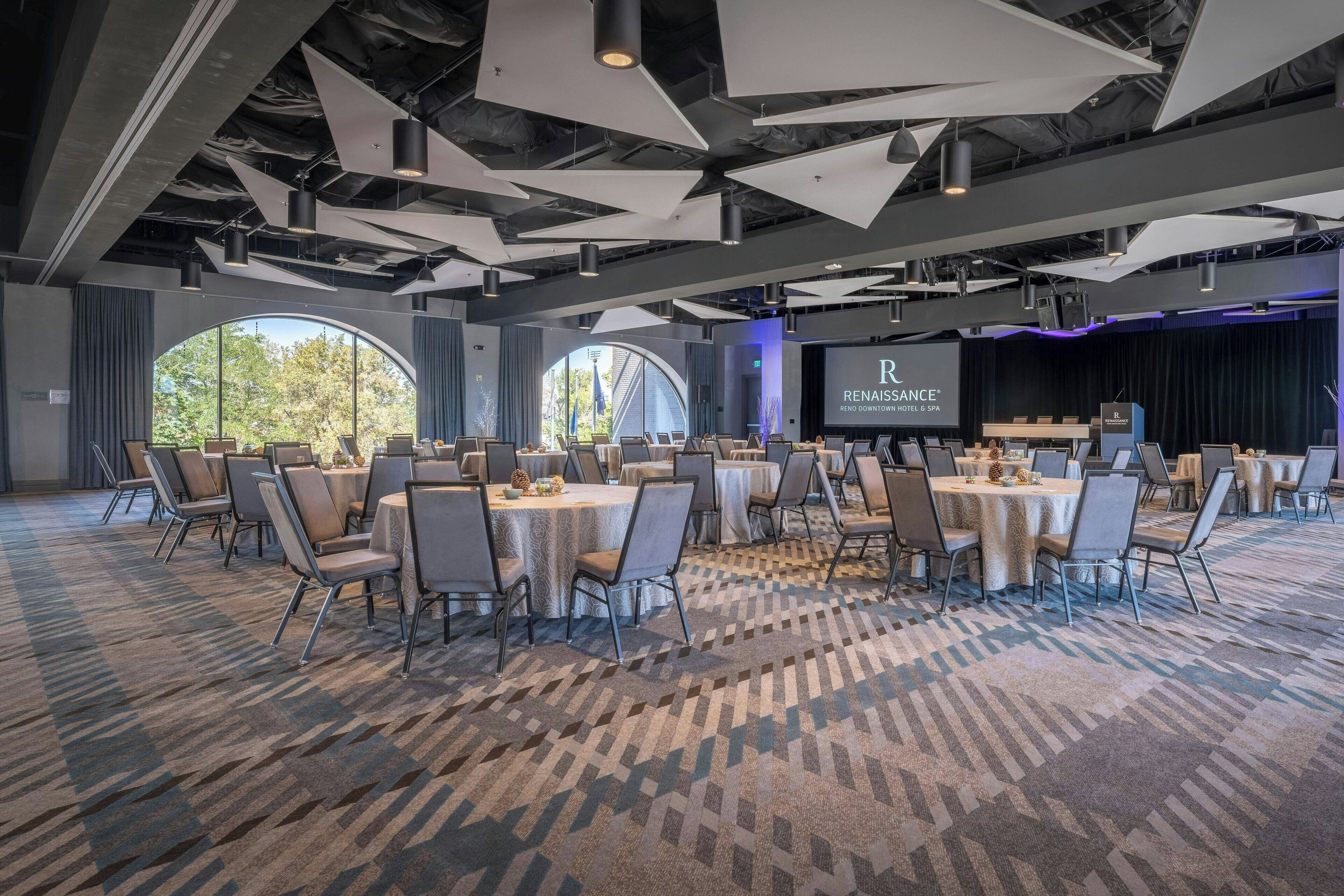 A conference room with round tables and chairs, large windows, and a stage with a screen displaying 'Renaissance Reno Downtown Hotel & Spa'.