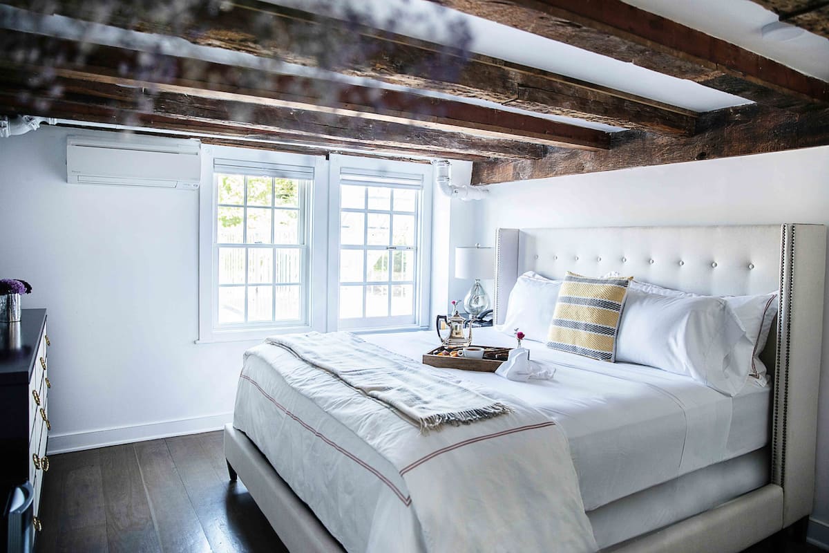 A bedroom with a large bed, wooden beams, a window, and a bedside table with a lamp.
