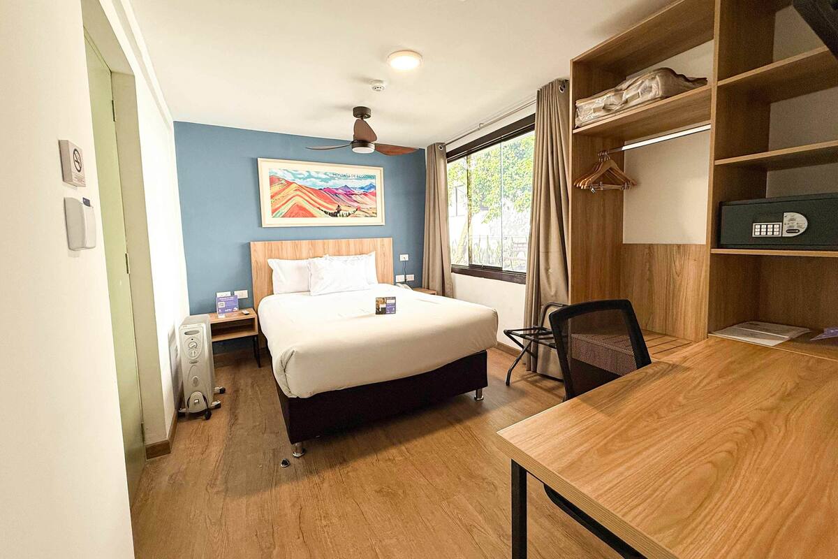 Une chambre d’hôtel comprenant un lit, un bureau, une chaise, une bibliothèque, une fenêtre avec des rideaux et un tableau au mur.