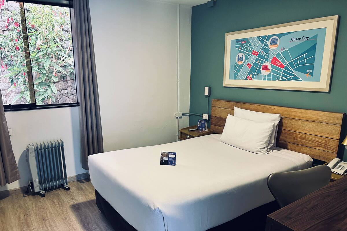 Une chambre d’hôtel avec un grand lit, une tête de lit en bois, une table de chevet avec une lampe, une carte encadrée accrochée au mur et un radiateur près de la fenêtre.