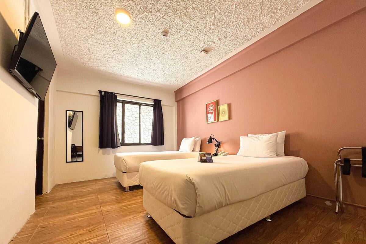Une chambre d’hôtel avec deux lits, un téléviseur à écran plat fixé au mur, une fenêtre avec des rideaux et un plancher en bois.
