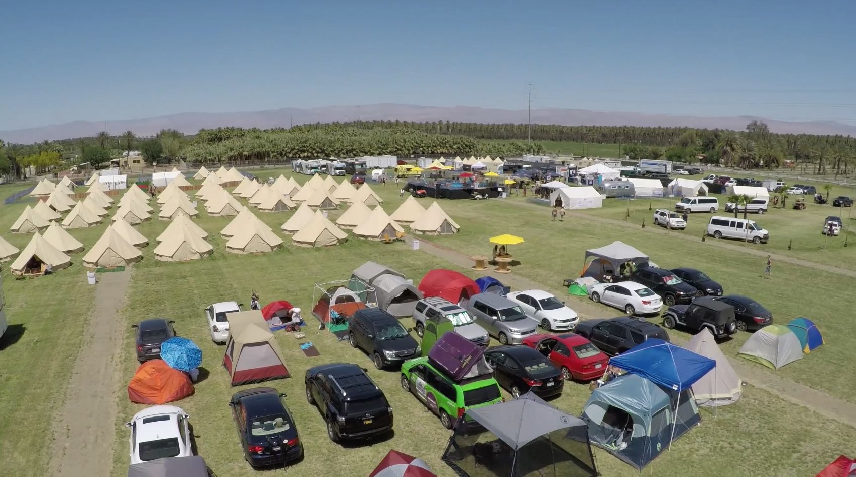 Um camping com várias tendas e veículos recreativos estacionados em campos gramados, com montanhas ao fundo.