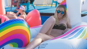 A person lounging on a colorful inflatable float in a pool area with a white fence and chairs in the background.
