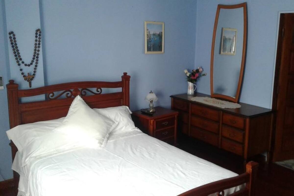 Une chambre à coucher avec un lit en bois, une commode, un miroir et un crucifix fixé au mur.