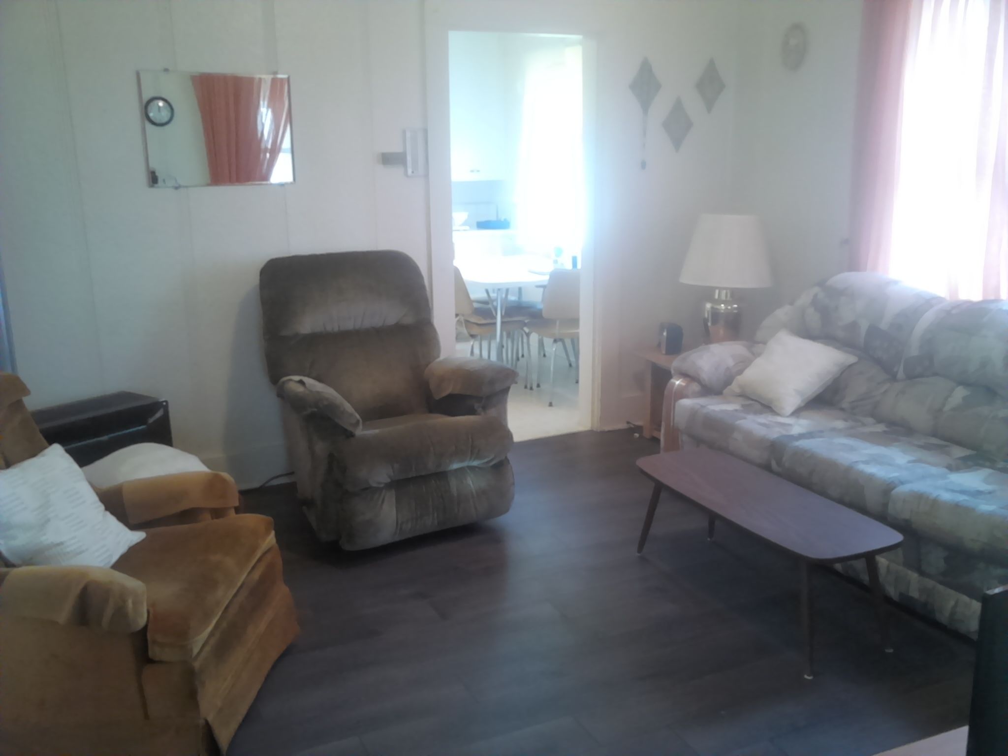 A living room with a brown armchair, a sofa, a small table, and a lamp. There is a door leading to a dining area.