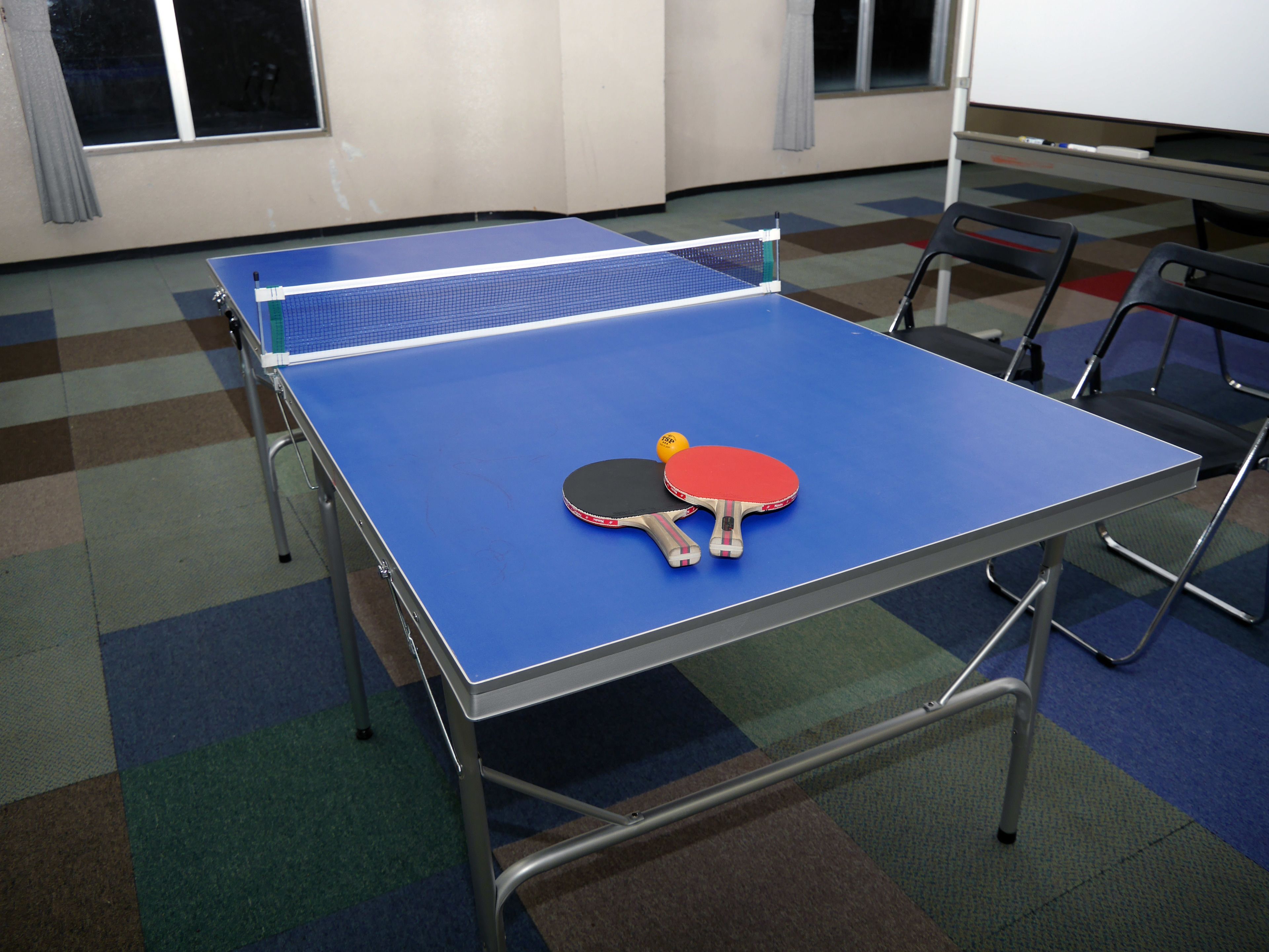 Uma mesa de pingue-pongue com duas raquetes e uma bola em um quarto com carpete estampado e cadeiras.