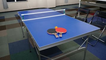 Uma mesa de pingue-pongue com duas raquetes e uma bola em um quarto com carpete estampado e cadeiras.