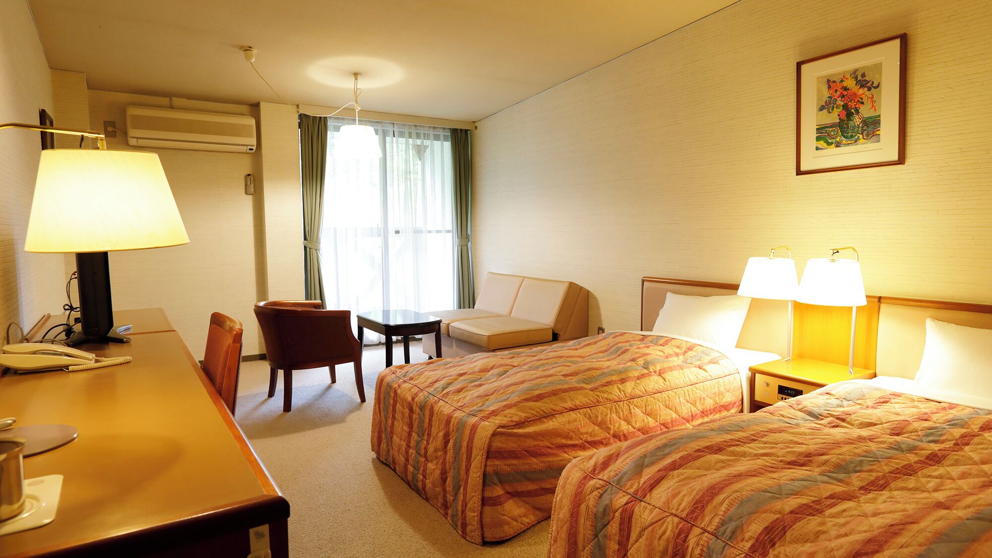 Ein Hotelzimmer mit zwei Betten, einem Schreibtisch mit Lampe, einem Sessel, einem Tisch und einem Fenster mit Vorhängen.