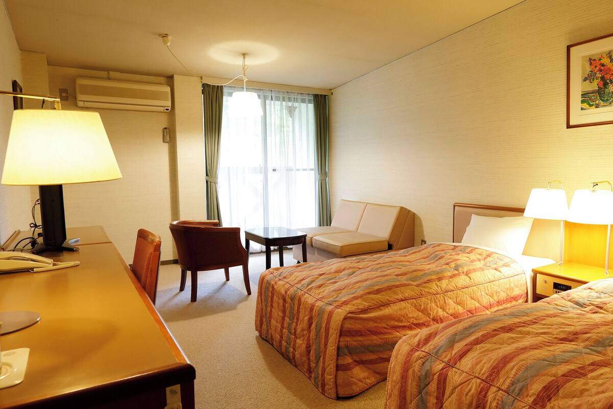 Une chambre d’hôtel avec deux lits, un bureau avec une lampe, une chaise, une table et une fenêtre avec des rideaux.
