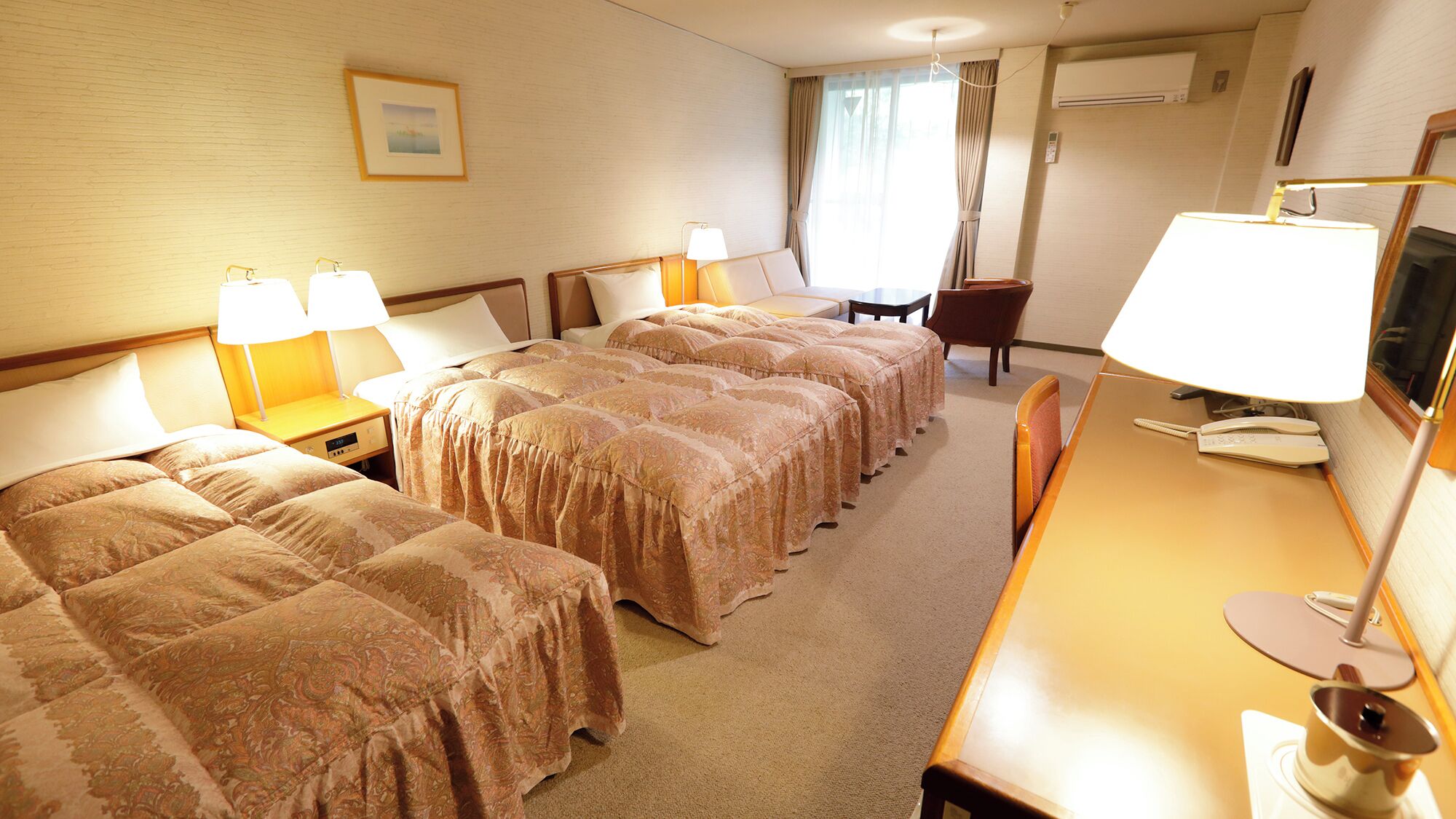 Ein Hotelzimmer mit zwei Betten, einem Schreibtisch mit Lampe, einem Sessel, einem Fernseher und einem Fenster mit Vorhängen.