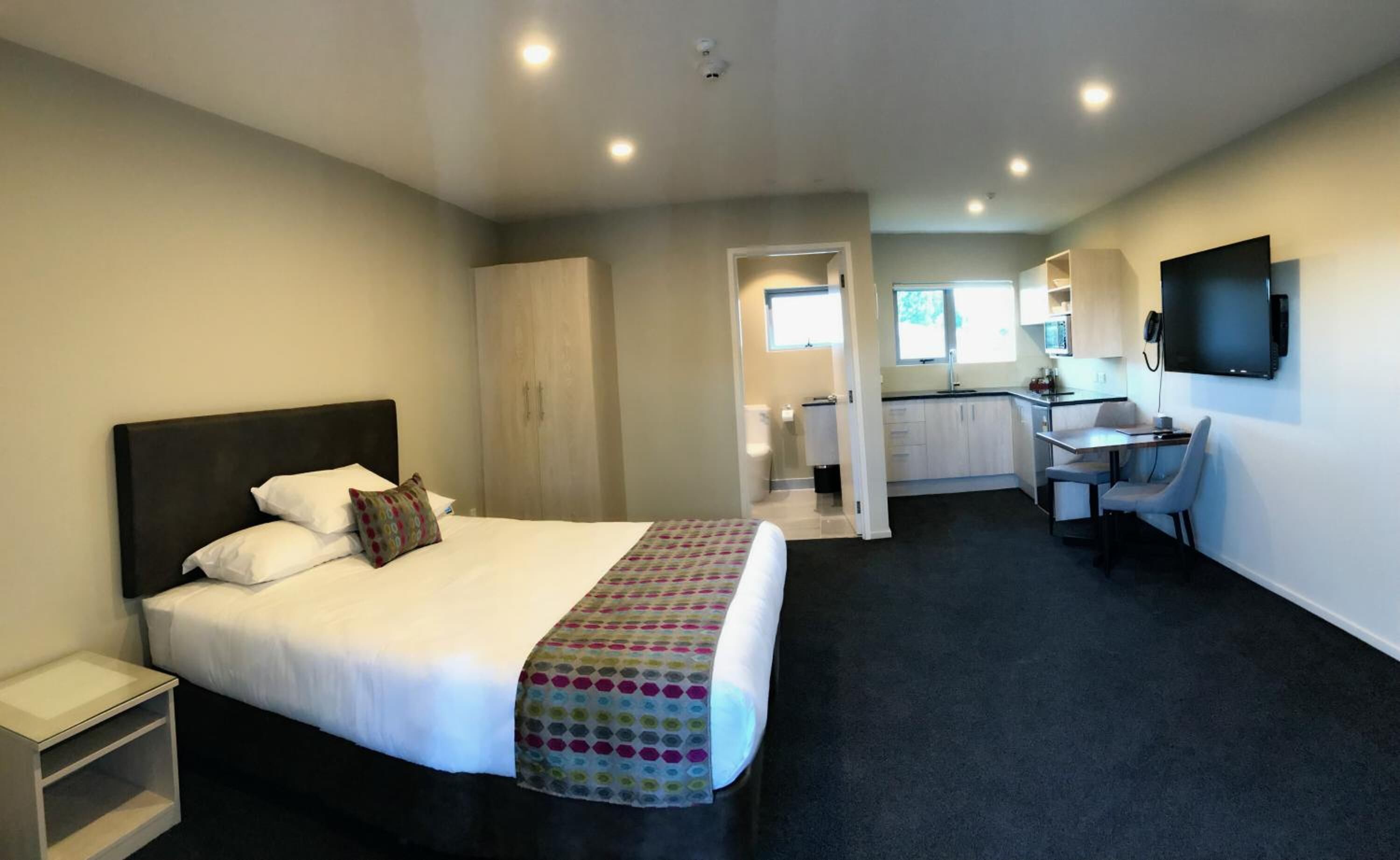 Habitación de hotel con cama, escritorio, silla, televisor y baño visible a través de una puerta abierta.
